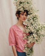 A person wearing a short-sleeved raspberry pink cotton voile blouse with a V-neckline, featuring short, slightly blousante sleeves and embroidered foliage and floral scroll embroidery.